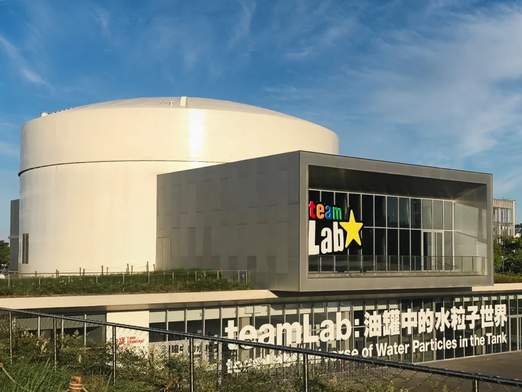 Exterior of a teamLab exhibition building featuring a large white cylindrical structure and a glass facade with the teamLab logo.