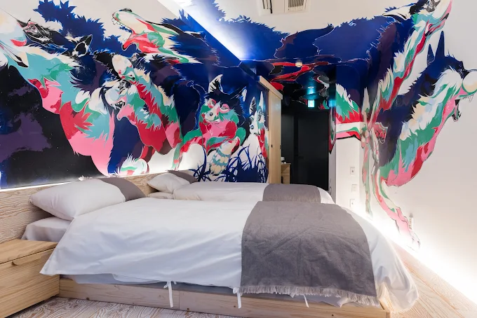 Interior of an artist-designed room at BnA Hotel Koenji, featuring two beds surrounded by a vibrant, floor-to-ceiling painted mural of wolves in bold pink, blue, and green colors.
