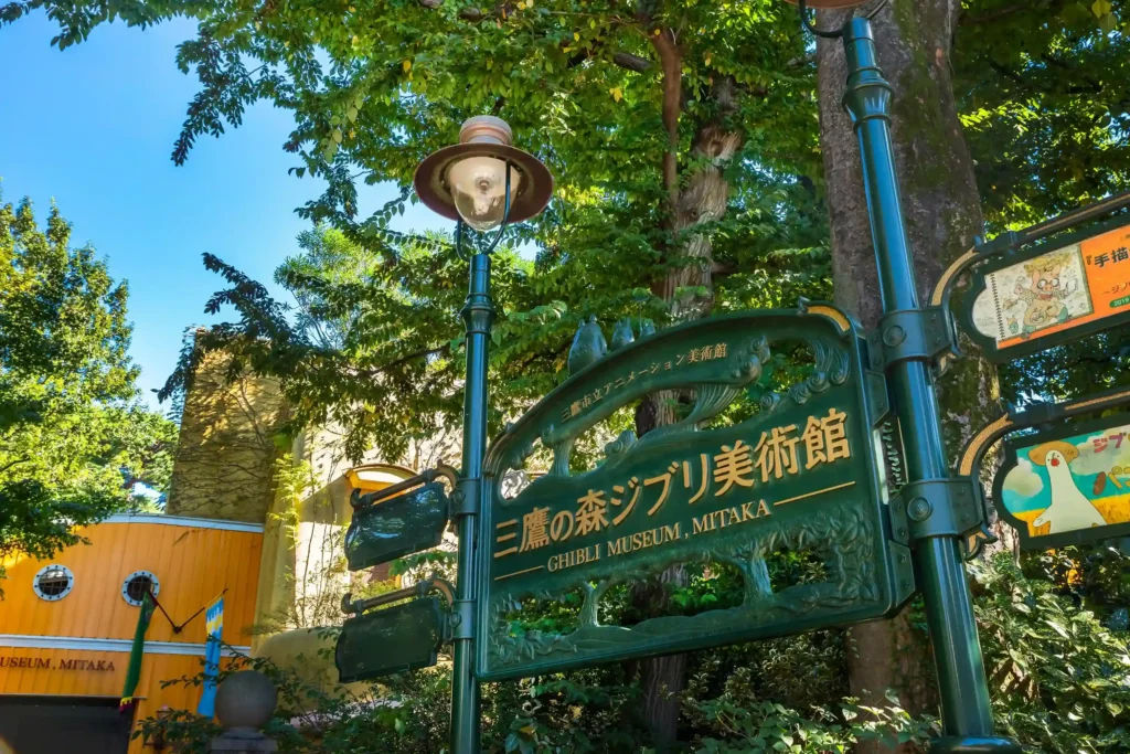 Ornate green entrance sign for the Ghibli Museum in Mitaka featuring a small Totoro sculpture, with colorful museum buildings in the background.