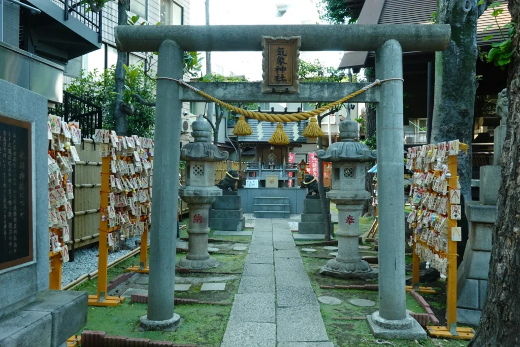 Paved path passing through a stone torii gate, framed by lanterns and racks filled with wooden prayer plaques (ema), leading to Kisho Shrine near Koenji Station.