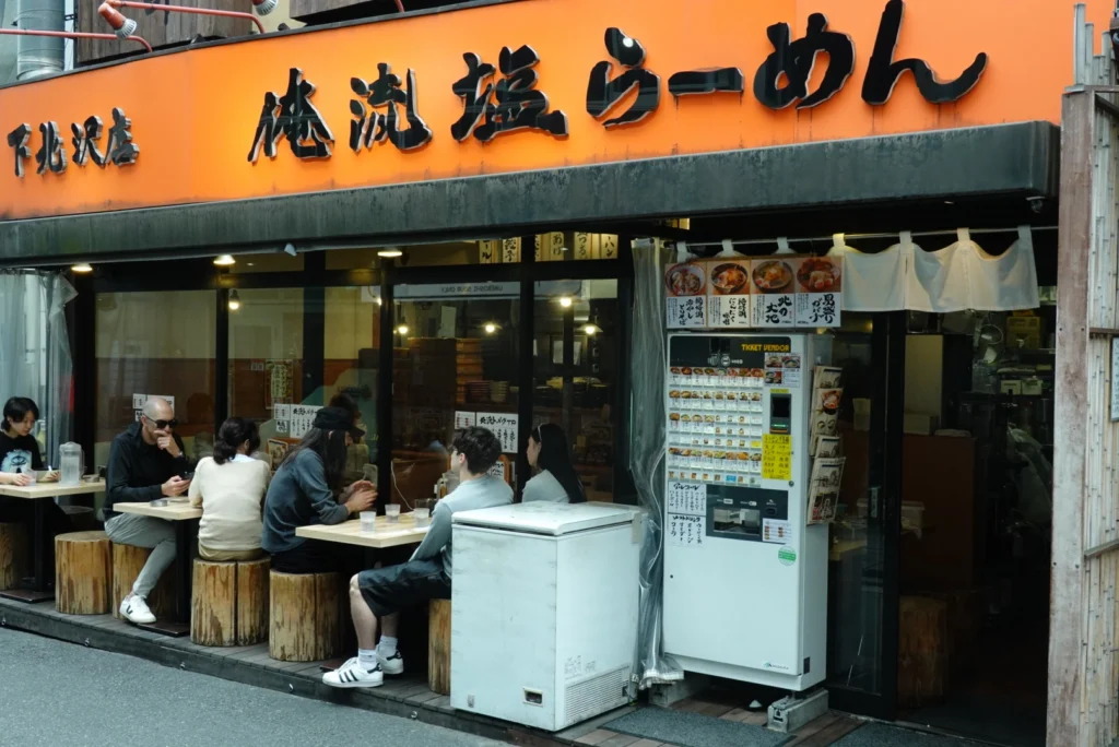 Diners eating at outdoor wooden tables in front of a traditional ramen shop in Shimokitazawa.