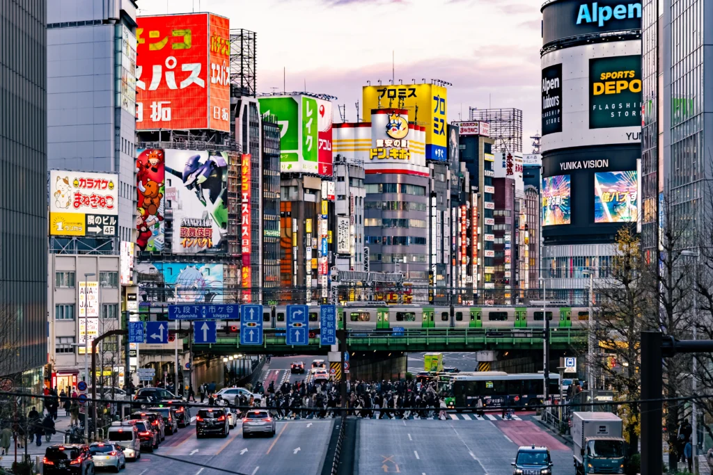 A bustling multi-lane street in Shinjuku at dusk, filled with cars and pedestrians crossing, framed by towering buildings covered in bright neon signs and a green JR Yamanote train passing on an elevated track.