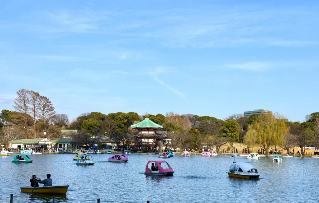 People riding colorful swan boats and rowboats on Shinobazu Pond in Ueno Park, with the green-roofed Bentendo Temple visible in the background.
