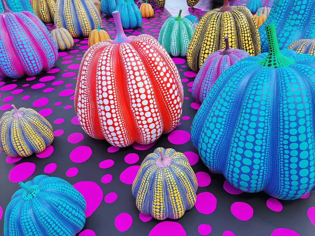 Vibrant, multi-colored polka dot pumpkin sculptures by contemporary artist Yayoi Kusama displayed on a dark surface with pink circles.